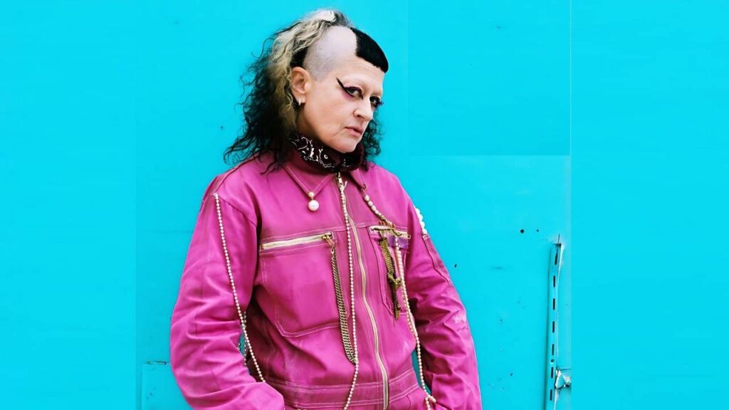 Erika Lust with a black-and-blonde mohawk in a pink jacket with pearls, posed against a bright turquoise wall.