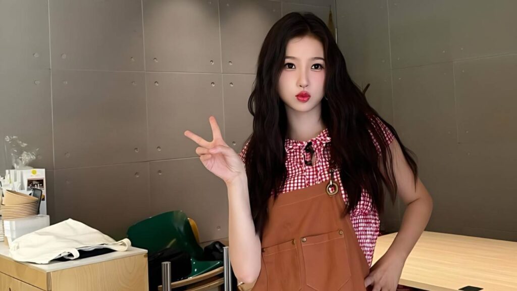 Esther Yu with long dark hair in brown overalls over a red gingham top, flashing a peace sign and puckering lips indoors.