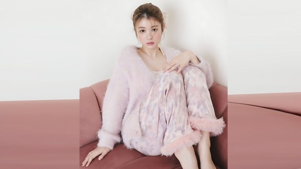 Fumika Baba with light makeup sits on a pink sofa, wearing a fluffy pastel cardigan and tie-dye pants, looking at the camera.