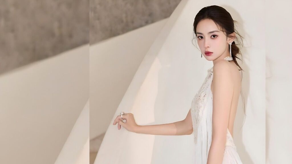 Gülnezer Bextiyar in a backless white gown poses along a white backdrop, with hand on a white railing and long earrings.