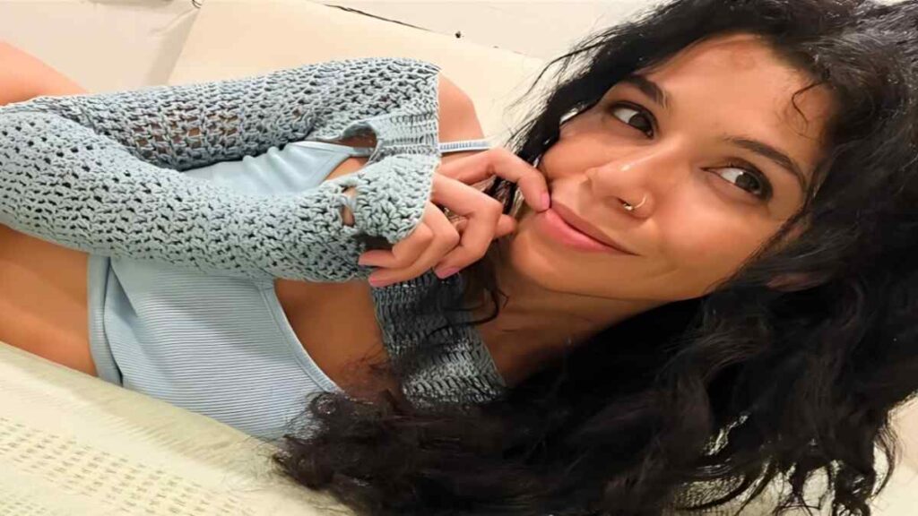 Harley Haze with long dark curls rests on a bed, chin on her hand, wearing a blue ribbed top and blue crochet sleeves.