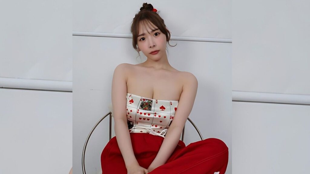 Himeka Iori on a metal chair, strapless card-print top and red pants, hair in a bun with a red clip, against a plain wall.