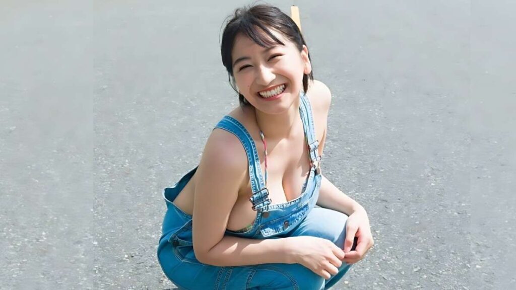 Ina Enohara with short dark hair in blue denim overalls crouching on a gravel surface under sunny light.