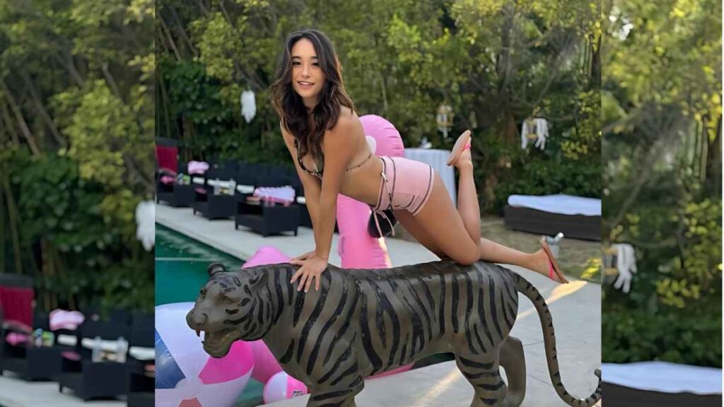 Isabella Nice in a pink bikini kneels on a tiger statue by a pool, smiling, with pink flamingo float and lounge chairs nearby.