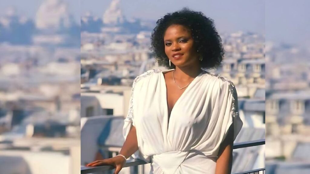 Jeannie Pepper with curly black hair in a white draped dress stands at a balcony railing, smiling over a sunlit cityscape.