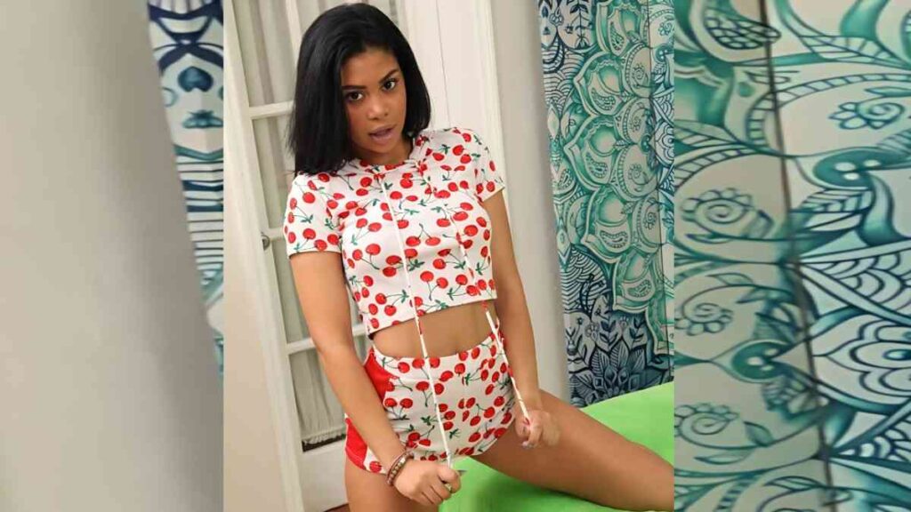 Jeni Angel in a white cherry-print crop top and matching shorts poses indoors by a doorway with turquoise patterned curtains.
