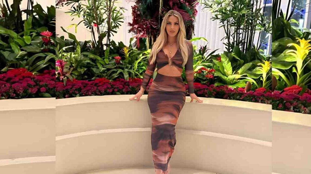 Jillisa Lynn in a brown tie-dye two-piece outfit with a cutout midriff stands on a curved white bench amid lush tropical plants and red flowers