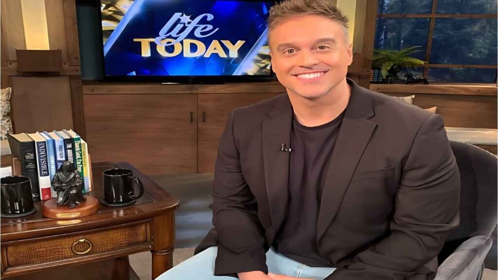 Joshua Broome in a black blazer sits on a studio couch beside a wooden table with books and mugs, Life Today on screen behind.