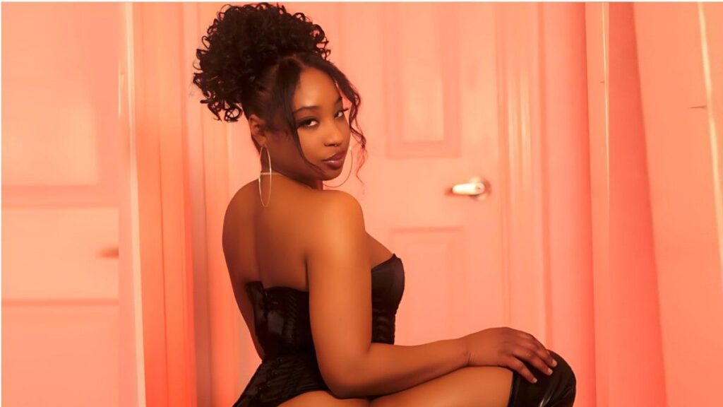 Lala Ivey in a black strapless corset, seated sideways against pink walls with an updo and hoop earrings.