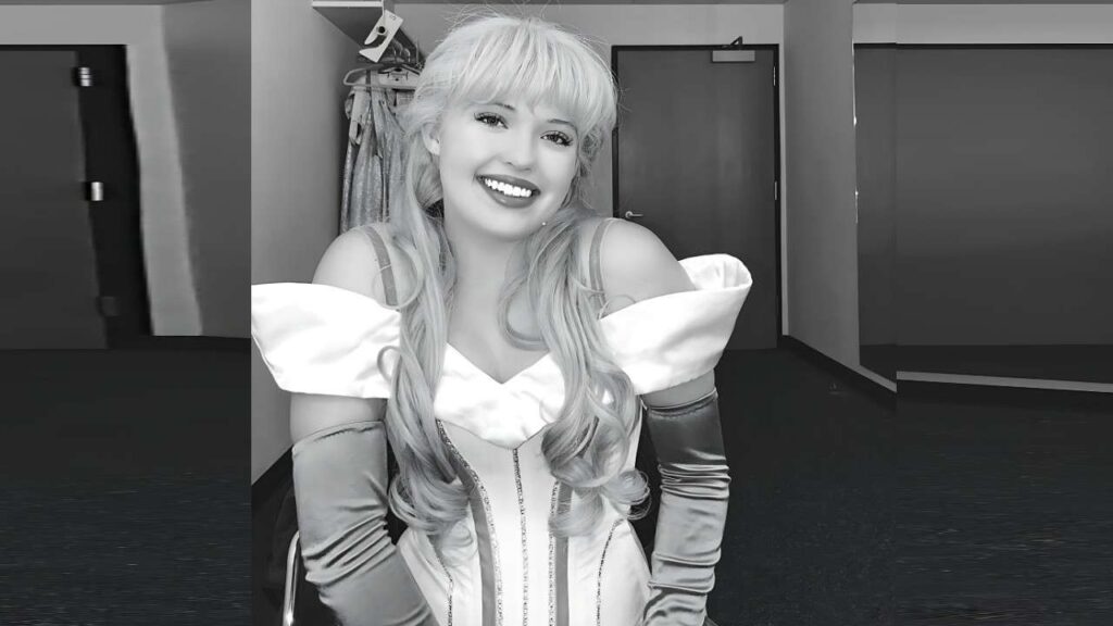 Lauren Taylor in a white off-shoulder fantasy gown with long satin gloves, seated in a backstage dressing room.