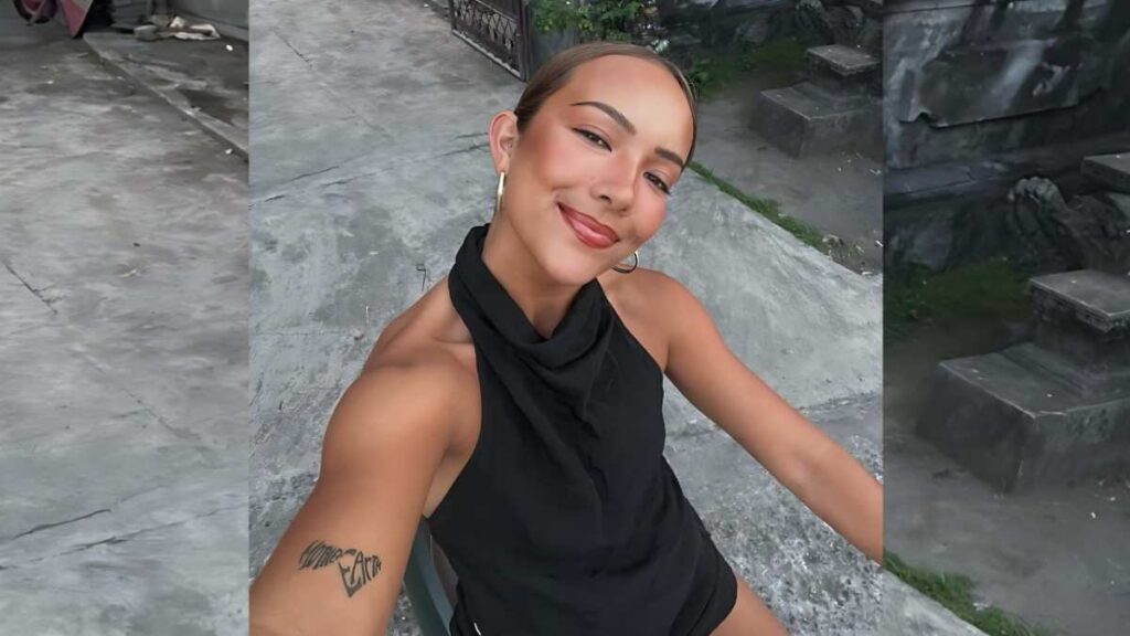 Leah Winters on gray stone steps, wearing a black halter top and gold hoops, taking a selfie with forearm tattoo visible.