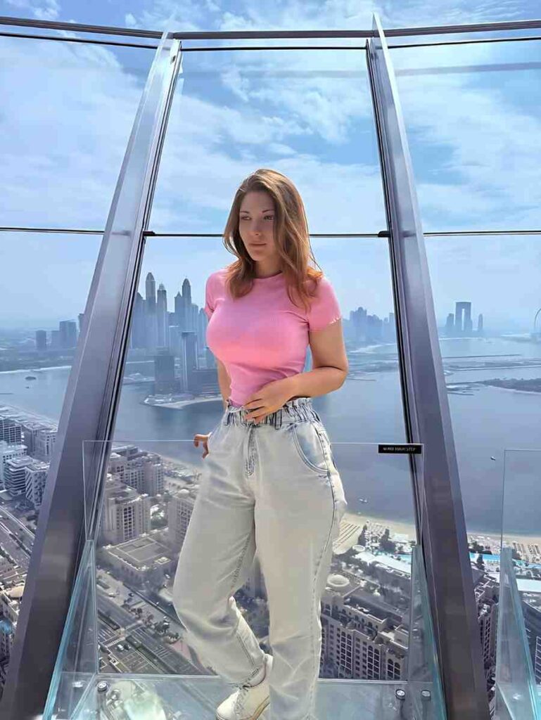 Lika Busy in pink tee and light jeans on a glass observation deck with a city skyline and river beneath a blue sky.