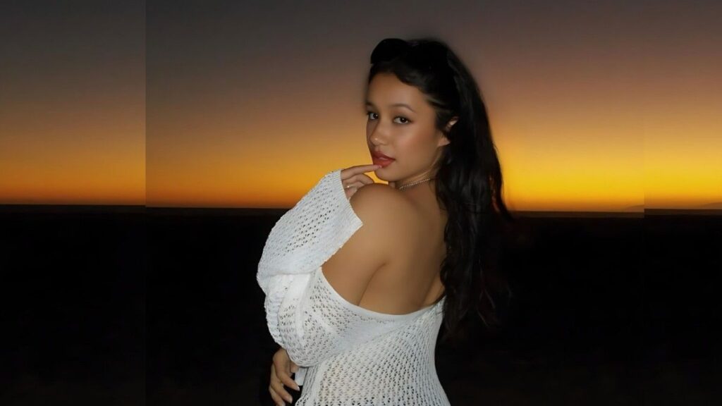 Lilith Grace with long dark hair in a white knit off-shoulder top, posing against a sunset gradient, finger to lips.