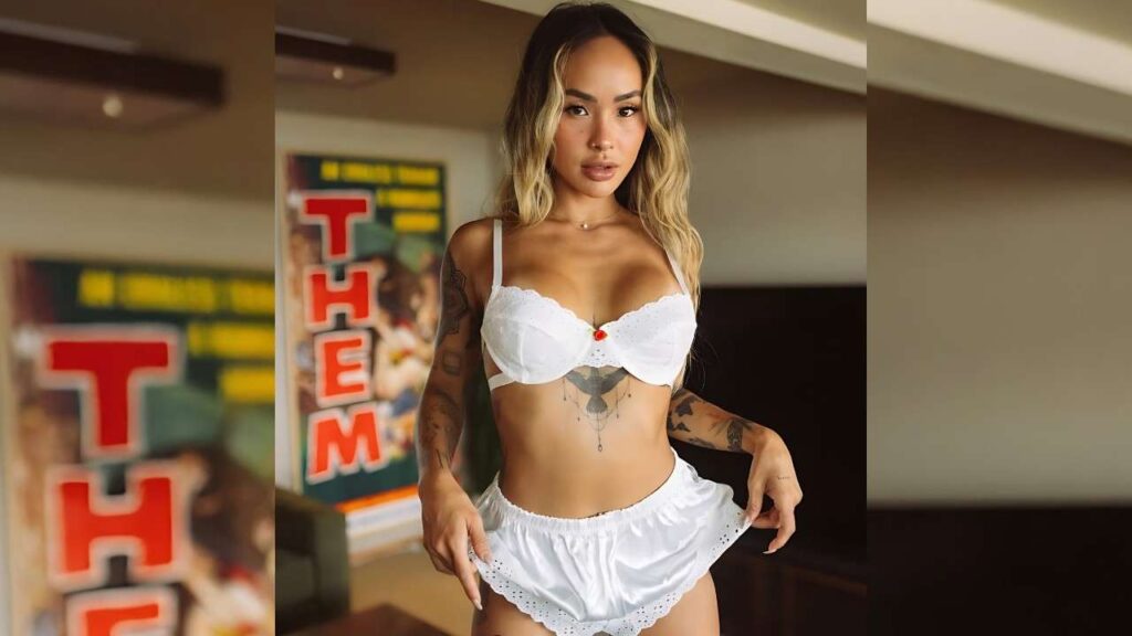 Linda Lan in white bra and shorts, blonde hair, tattoos on arms and torso, indoors beside posters with red THEM.