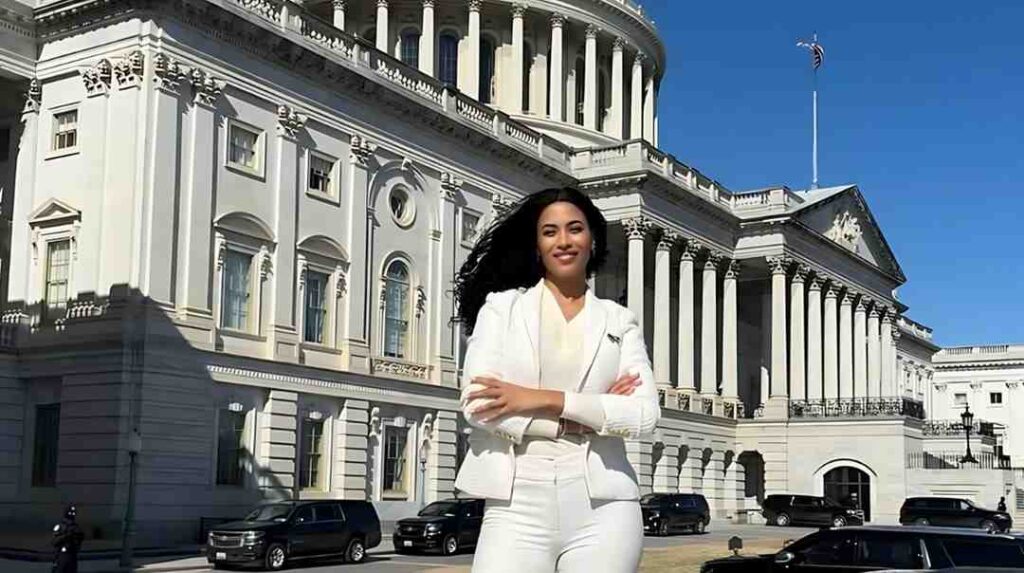 Lisa Phillips in a white suit stands with arms crossed in front of a grand neoclassical building under a blue sky.