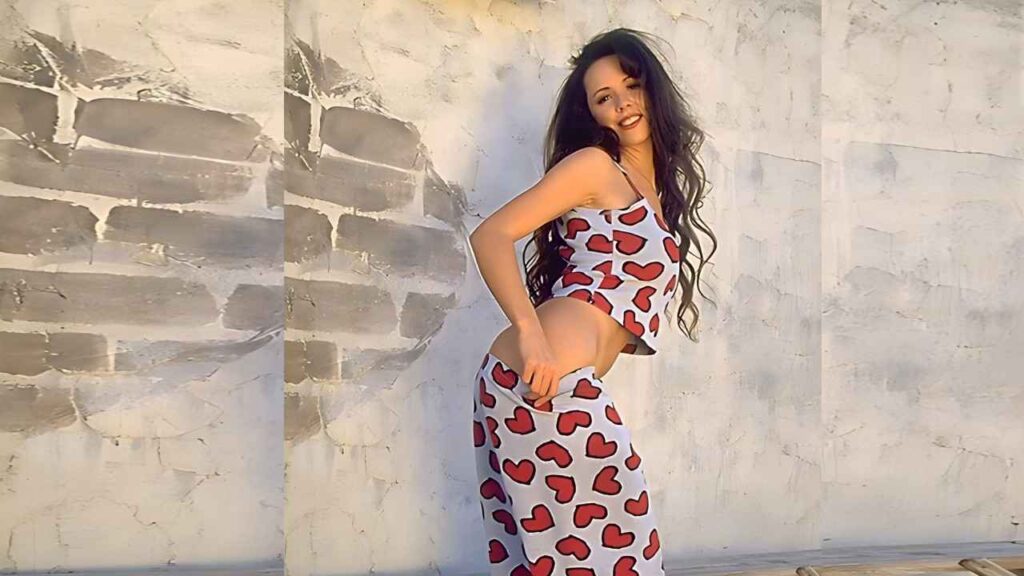 Lorissa McComas with long dark hair in a red heart print tank top and skirt poses against a sunlit, textured white wall, smiling.