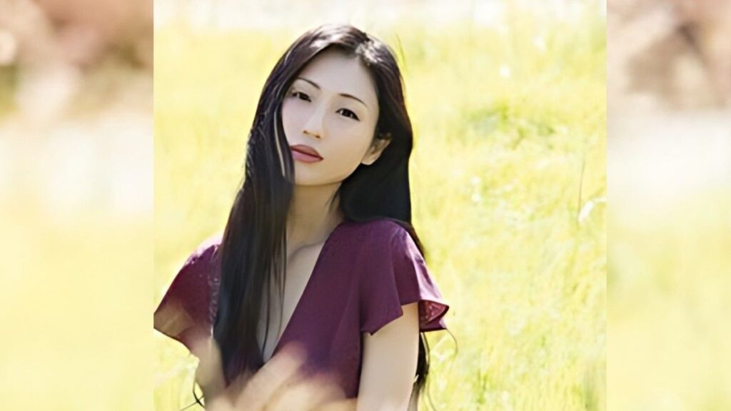 Mitsu Dan with long dark hair in a purple V-neck dress stands in a sunlit yellow field, gazing softly at the camera.