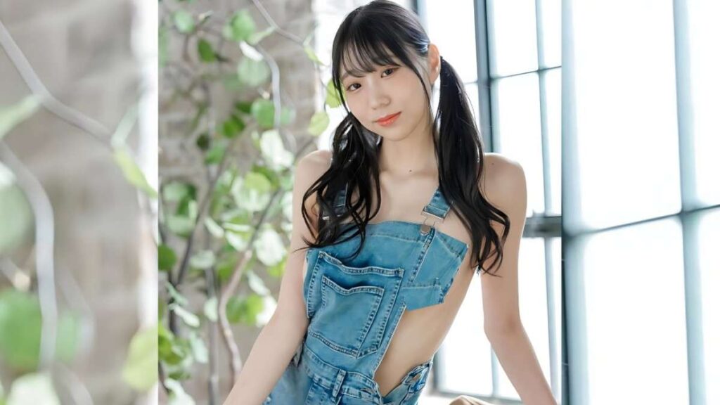 Mitsuki Momota in a denim one-strap romper, twin pigtails, posed near large grid windows with greenery.