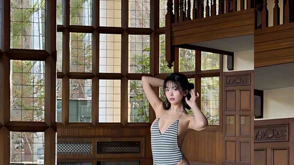 Moeka Sasaki in a gray striped swimsuit poses with hands in her hair inside a wood-paneled room with large grid windows.