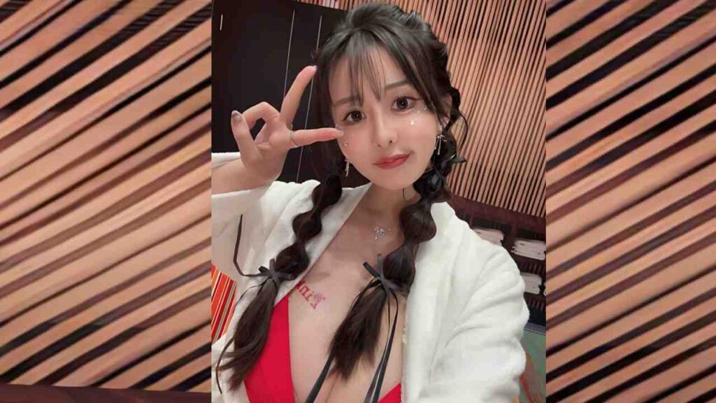 Momoka Kagura with two braided pigtails in a white robe over a red top, posing peace-sign selfie against striped wooden wall.