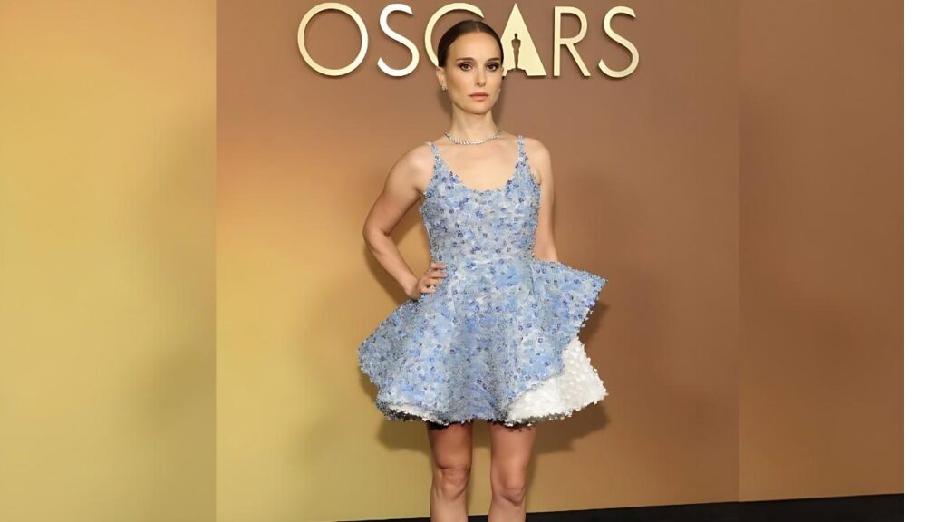 Natalie Portman in a pale blue floral dress with a voluminous layered skirt poses on a tan Oscars backdrop.