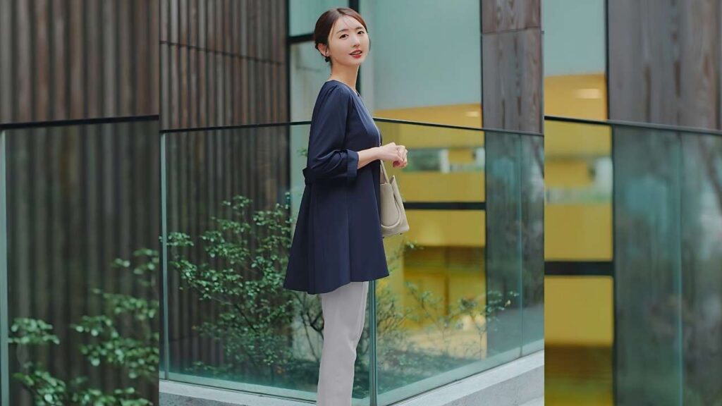 Nonoka Ono in navy tunic and light gray pants stands beside a glass storefront, holding a beige handbag and smiling.
