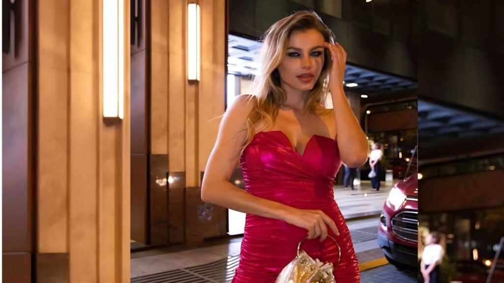 Olga De Mar in a hot-pink strapless dress poses outside an illuminated building, touching her hair and holding a clutch.