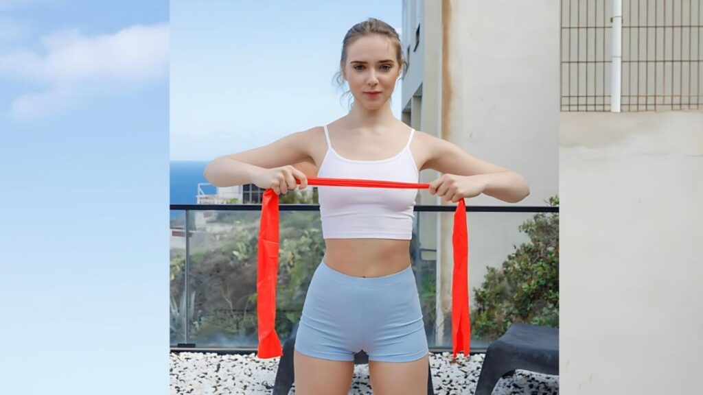Oxana Chic in a white sports bra and light blue shorts stretches a red resistance band across her chest on a balcony.