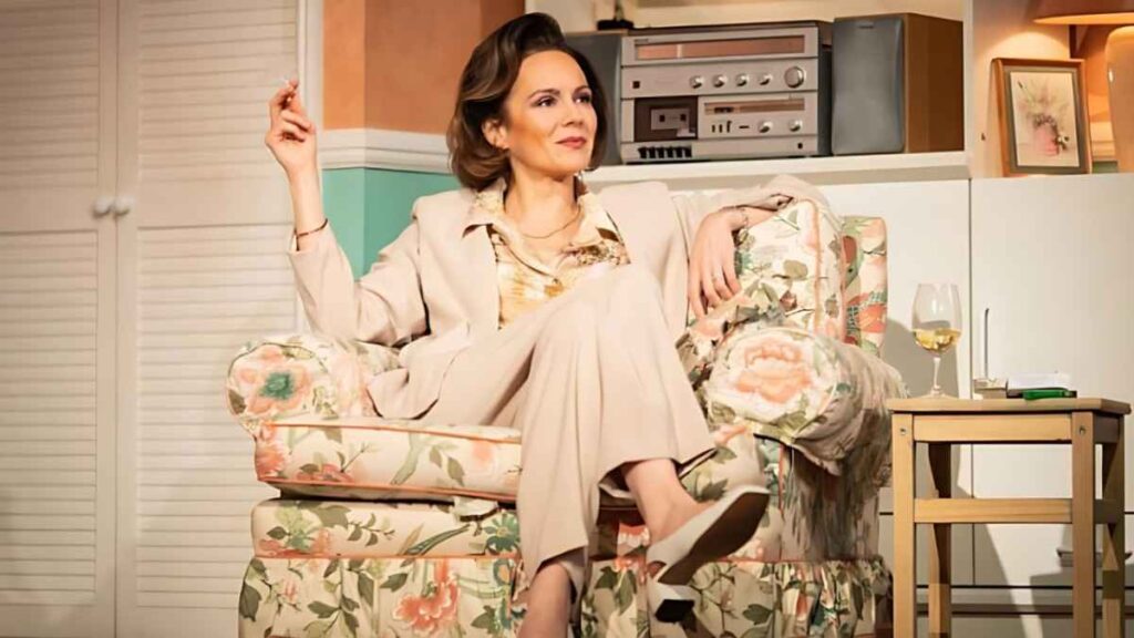 Rachael Stirling in a beige suit lounges in a floral armchair, cigarette raised, retro living room with a vintage stereo.