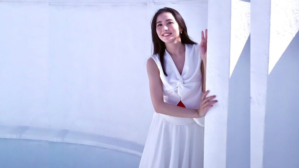 Ren Ishikawa in a white sleeveless wrap blouse and flowing skirt leans on a white pillar, smiling against a blue backdrop.