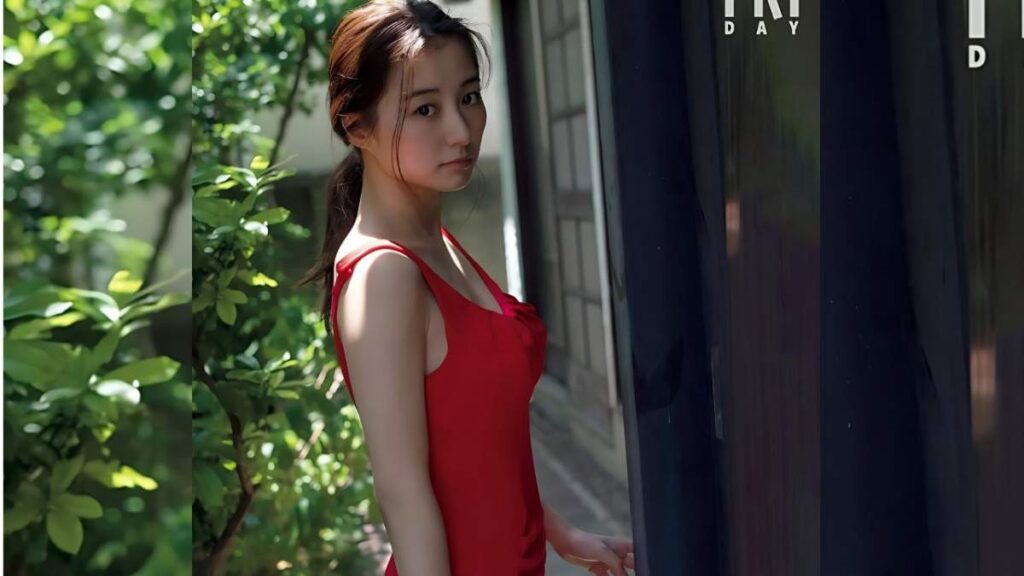Riho Takada in a red dress with thin straps stands near a dark glass wall, looking at the camera, greenery on the left.
