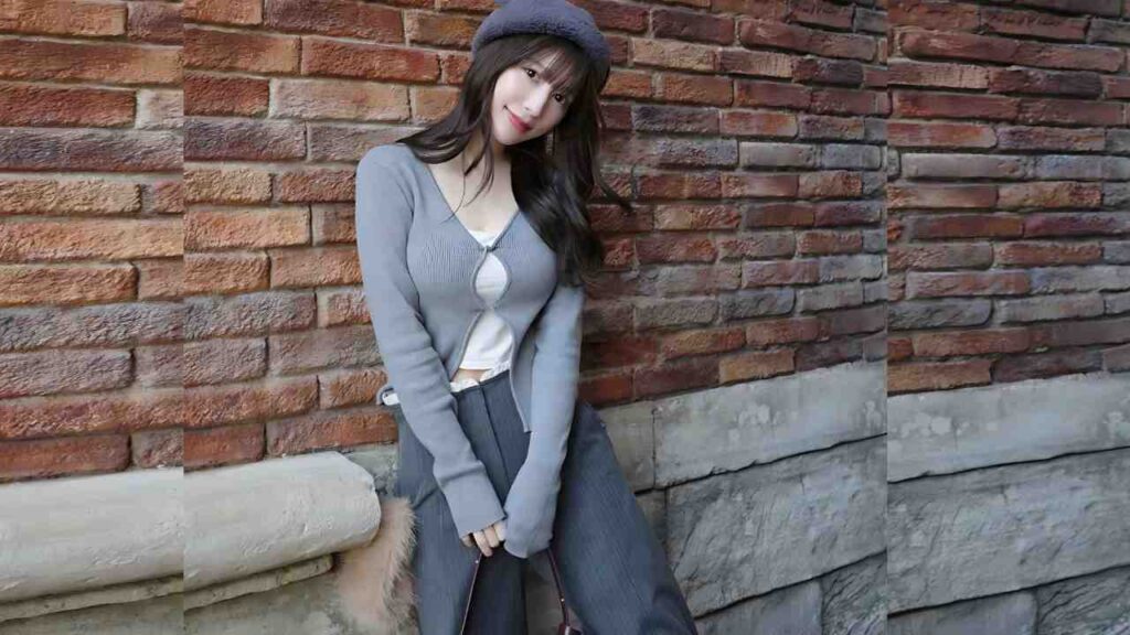 Risa Yukihira with long dark hair in a blue cardigan and beret, leaning against a red brick wall, smiling softly.