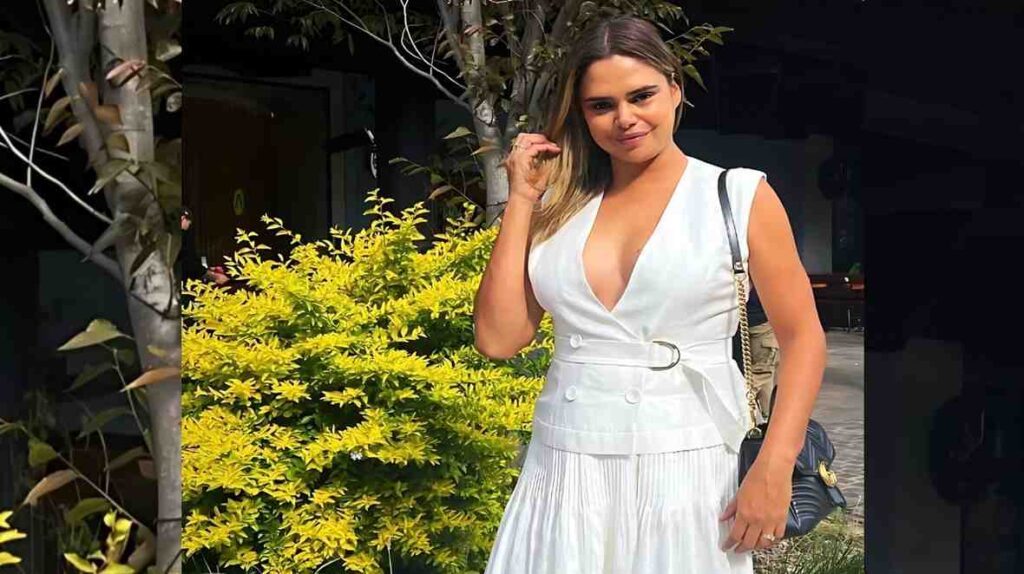 Samantha Harris in a white belted V-neck dress with a pleated skirt, touching her hair, carrying a black quilted bag.