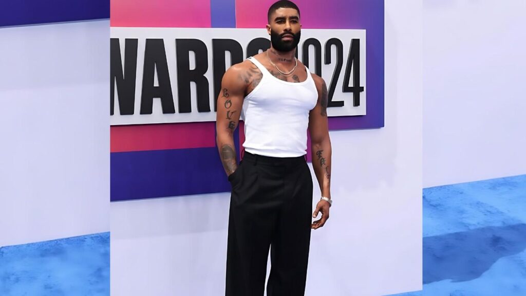 Skyy Black in a white tank top and black trousers stands with hands in pockets before a vibrant backdrop and tattoos.