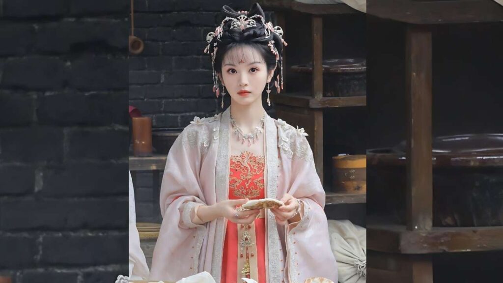 Tian Xiwei in pale pink hanfu with ornate silver headdress, holding a pastry in a rustic kitchen.