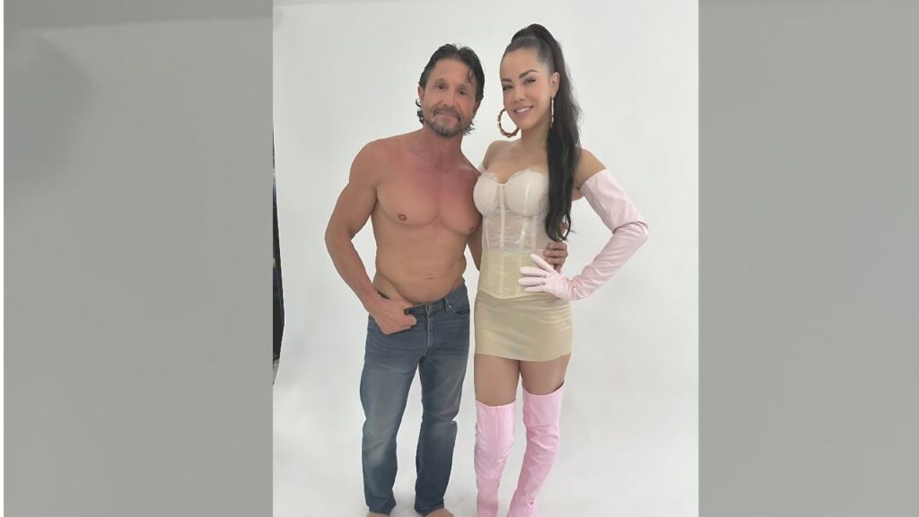 Tommy Gunn in jeans stands close with a smiling woman in beige corset and mini skirt, pink gloves and boots.