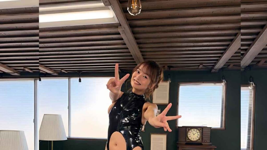 Tsukasa Wachi in a glossy black latex bodysuit poses with peace signs in a studio with exposed wooden beams, large windows, and a vintage clock.