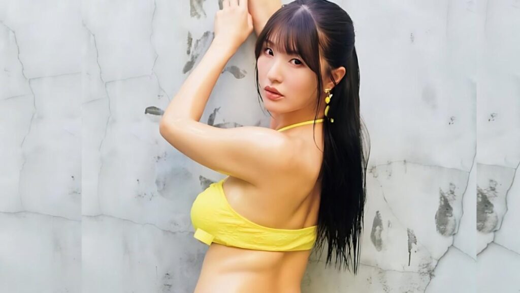 Tsumugi Hara in a yellow bikini top leans against a cracked white wall, arms raised, long dark hair, wearing yellow earrings.