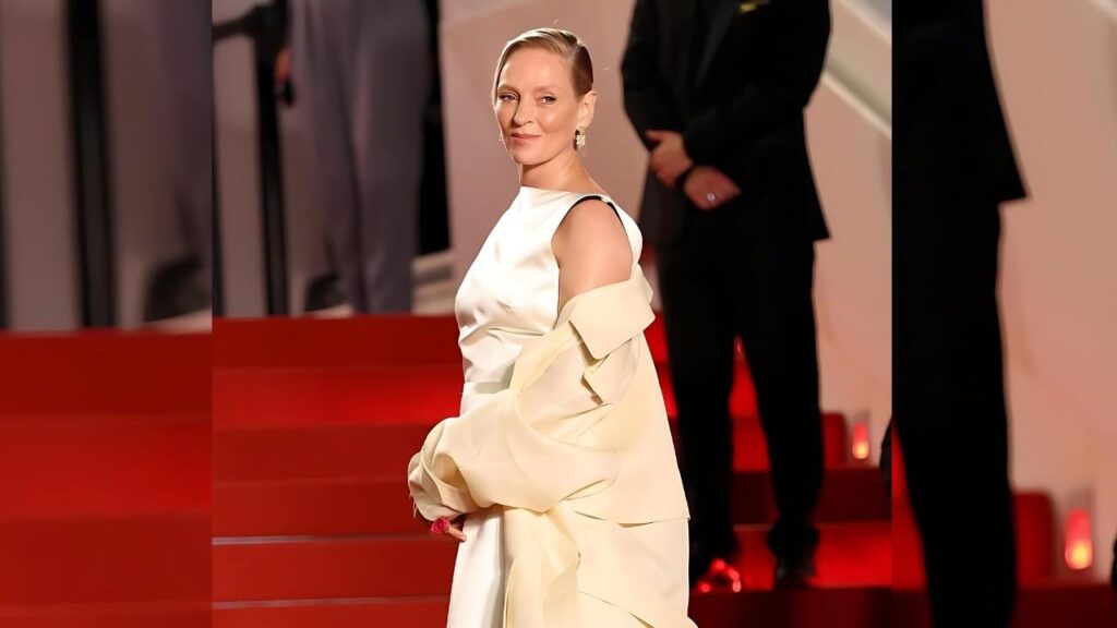 Uma Thurman in a white satin gown with a cream coat draped over her arms on a red carpet, blonde hair slicked back, smile.