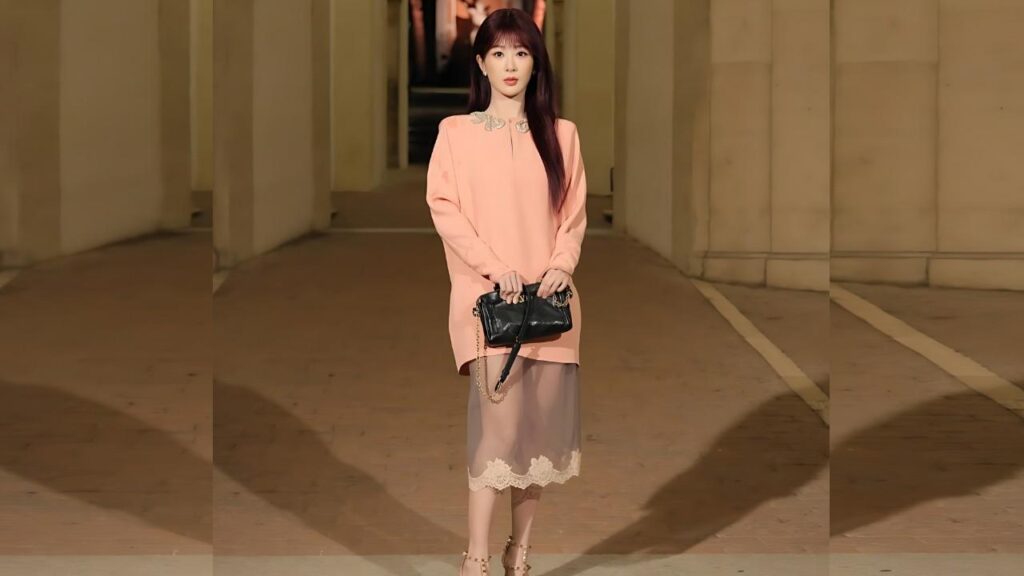 Yang Zi with long dark hair in a peach sweater and lace-hem skirt stands in a pillar-lined hallway, holding a small black chain-strap bag.