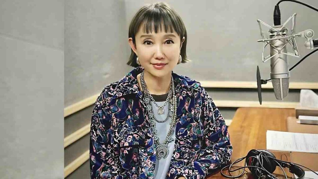 Youki Kudoh with short bob sits at a recording studio table by a large microphone, wearing a floral jacket and layered necklaces.