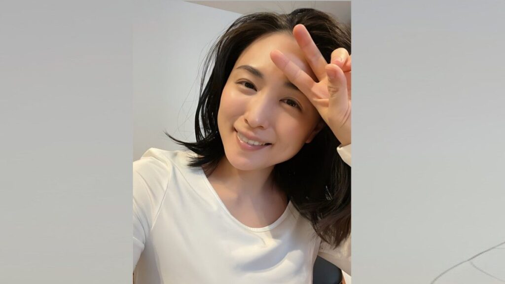 Yukie Kawamura with shoulder-length black hair in a white top, making a peace sign near her forehead indoors.