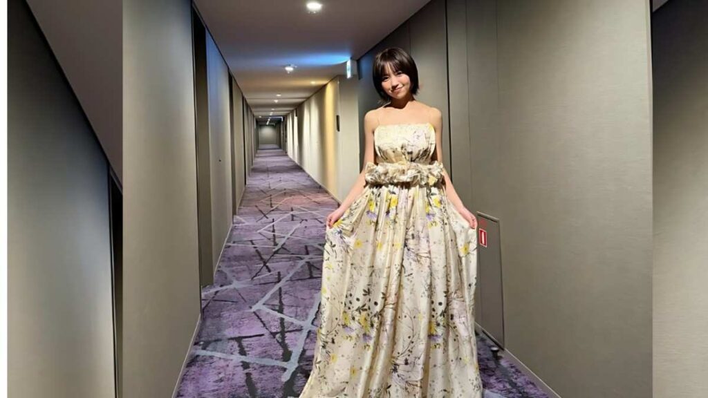Yuno Ohara in a strapless floral gown smiles in a beige hotel hallway, holding the skirt as corridor recedes behind her.