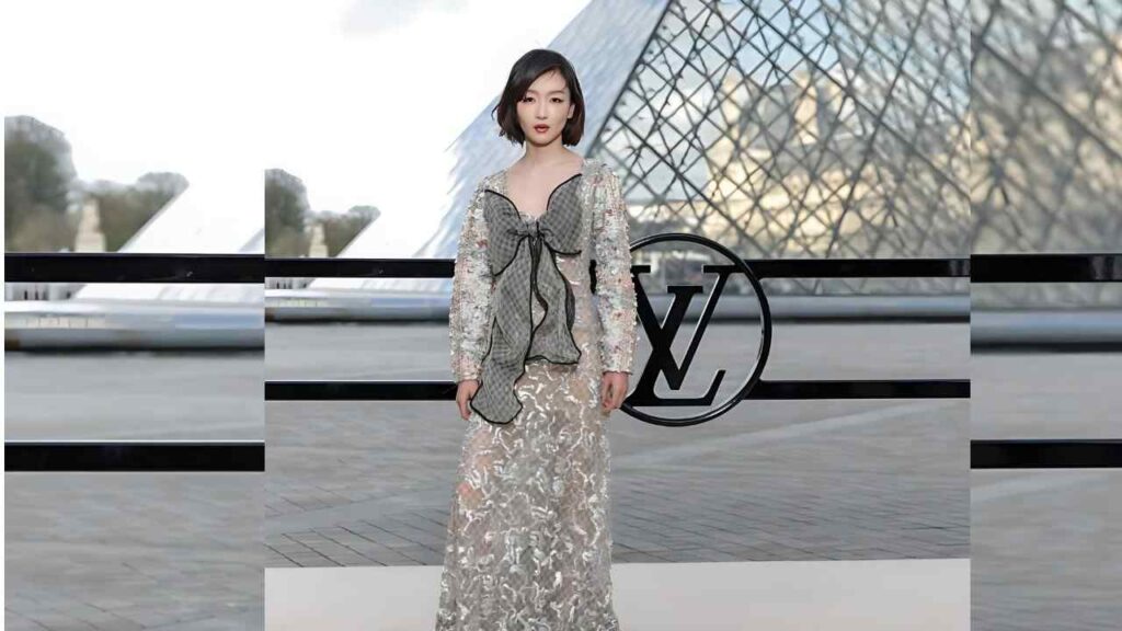 Zhou Dongyu with a short bob wears a pale sequined lace gown with a large bow, posing before a Louvre glass pyramid and LV logo.