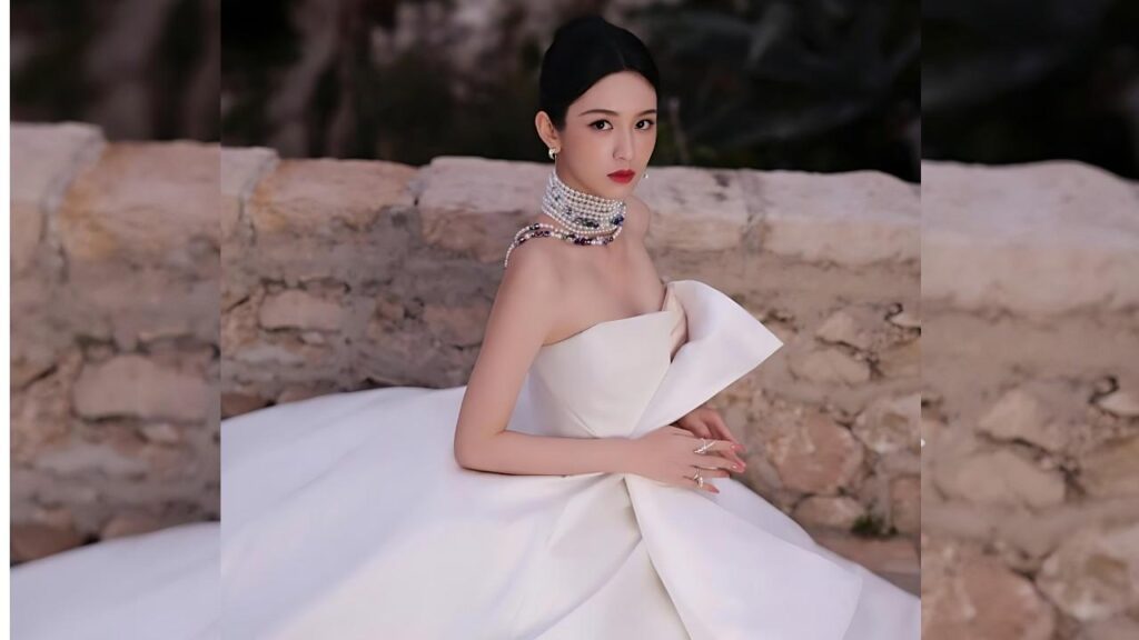 Zhou Ye in a strapless white gown with a bow sits by a stone wall, wearing pearl necklace and earrings, sleek hair.