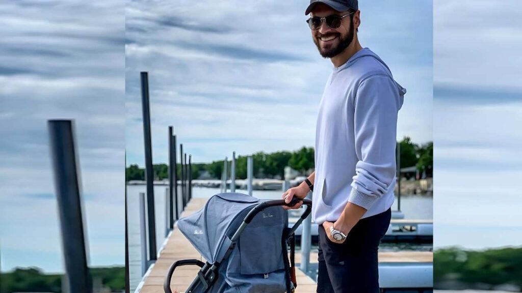 Adan Canto in a light gray hoodie, cap, and sunglasses pushes a blue baby stroller along a wooden dock beside calm water.