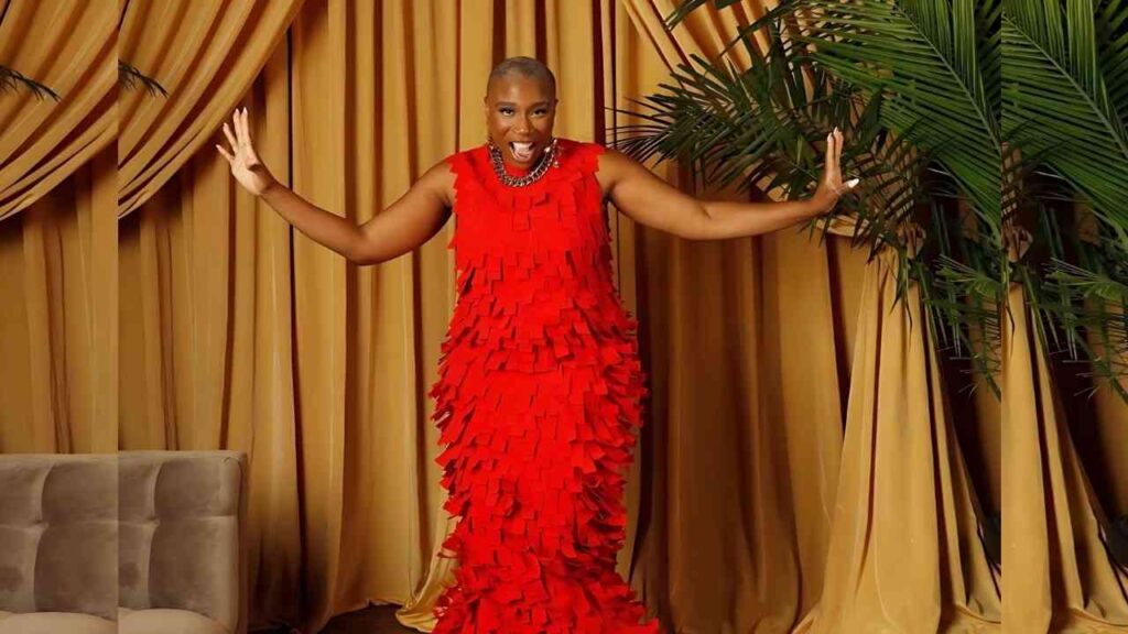 Aisha Hinds with a shaved head wears a bright red fringe dress, arms outstretched before gold curtains and lush plants.