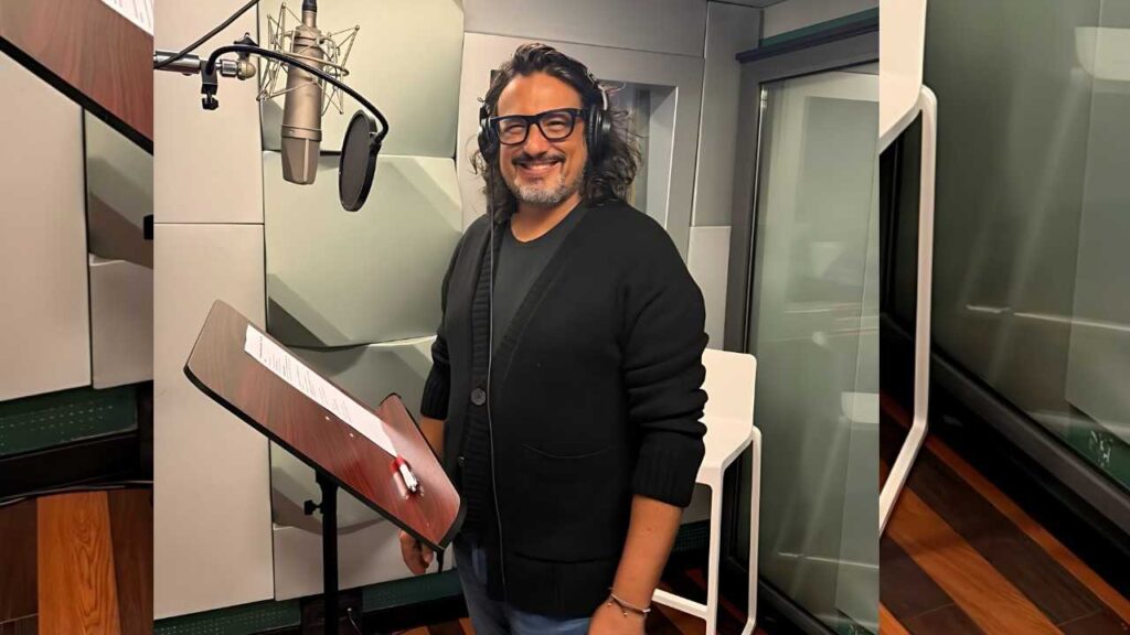 Alessandro Borghese with glasses and long hair, wearing headphones, stands at a music stand in a recording studio beside a mic with a pop filter.