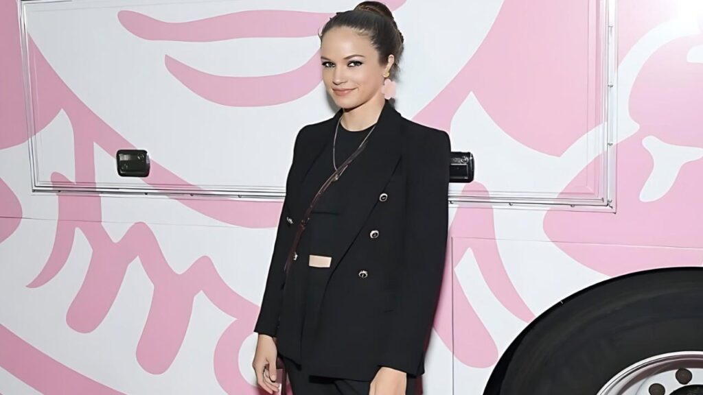 Alexis Knapp in a black blazer stands beside a pink and white vehicle, hair in a bun, pink earrings, subtle smile.