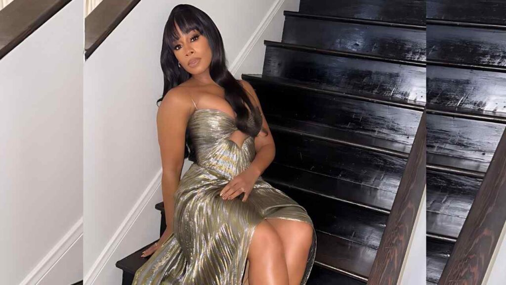 Anika Noni Rose in a metallic gold cut-out bodice, slit dress sits on a dark wooden staircase, with long black hair and bangs.